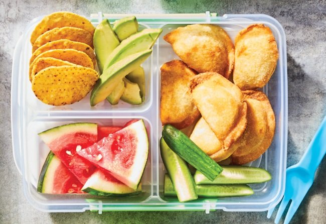 Plastic lunch container with compartments of chips, fruit, and empanadas on a gray surface.