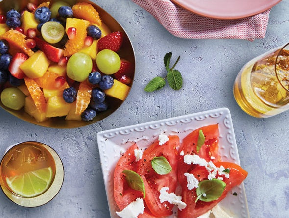 Fruit salad in a bowl and tomato dish with feta cheese on a light blue surface.