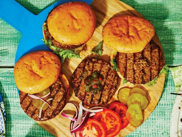 Three grilled burgers on a wooden board with vegetables on a green surface
