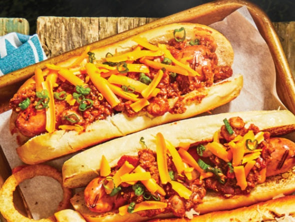 Two hot dogs with chili, cheese, and green onions on buns, served on a wooden tray.
