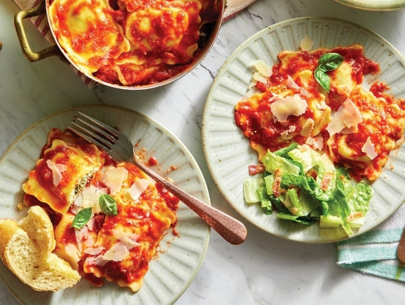 Two plates of ravioli with tomato sauce, cheese, and basil on a marble surface.