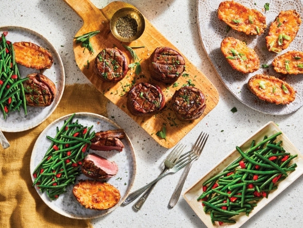 Dinner setting with plates of food including green beans, roasted potatoes, and steaks on a textured surface.
