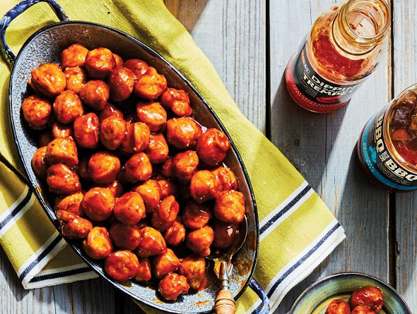 Baked frozen meatballs in a baking dish with a bottle of hot sauce on a wooden table.
